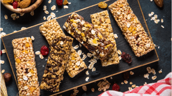 A black rectangular plate holds a collection of different favored nutrition bars beside a bowl of nuts and berries with oats sprinkled on top and the table around them.